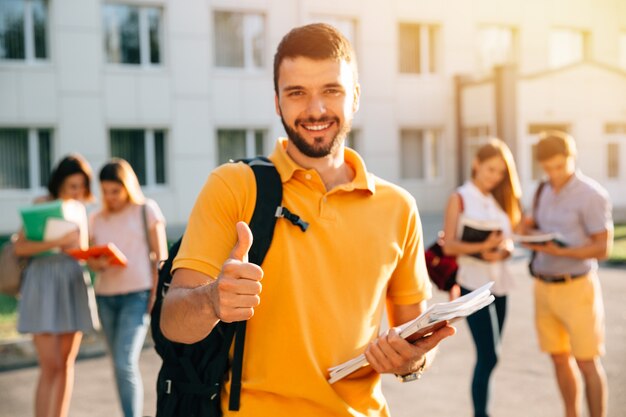 Estudante de sorriso atrativo novo que mostra o polegar acima fora no terreno na universidade.