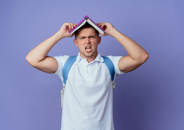 Estudante do sexo masculino bonito e descontente com uma bolsa nas costas coberta com um livro isolado no azul