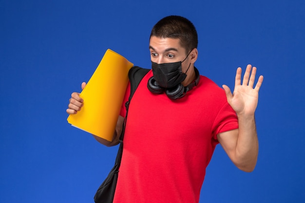 Estudante do sexo masculino com vista frontal, camiseta vermelha, máscara e mochila segurando uma pasta amarela na mesa azul.