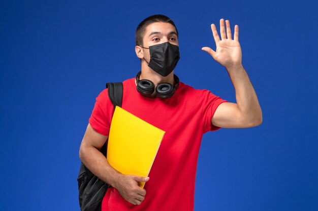 Estudante do sexo masculino de vista frontal em t-shirt vermelha usando máscara com mochila segurando arquivo amarelo sobre o fundo azul.