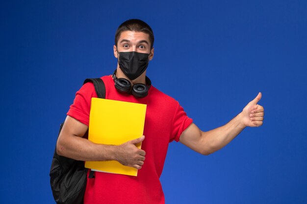 Estudante do sexo masculino de vista frontal em t-shirt vermelha usando máscara com mochila segurando arquivo amarelo sobre o fundo azul.