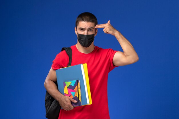 Estudante do sexo masculino de vista frontal em t-shirt vermelha, usando mochila em máscara estéril preta segurando arquivos posando sobre fundo azul.