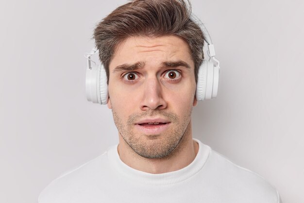 Estudante do sexo masculino preocupado intrigado com cabelos escuros ouve palestra via fones de ouvido tenta lembrar informações para o exame parece incomodado vestido casualmente isolado sobre fundo branco. Reações humanas