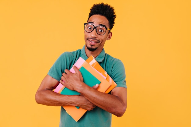 Estudante encaracolado espantado fazendo caretas engraçadas. jovem inteligente de óculos segurando livros.