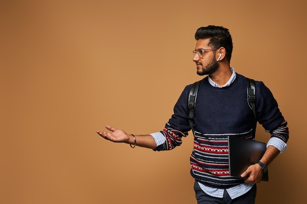 Foto grátis estudante indiano bonito elegante com laptop e mochila apontando a mão para a parede.