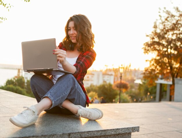 Estudante jovem alegre sentado com laptop ao ar livre no início da manhã ao nascer do sol Foto Premium