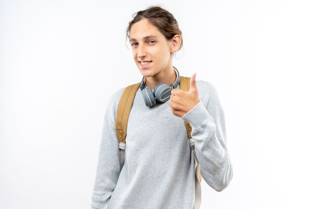 Estudante jovem sorridente usando mochila com fones de ouvido no pescoço aparecendo o polegar