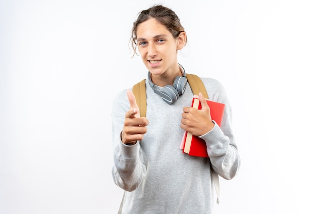 Foto grátis estudante jovem sorridente usando mochila com fones de ouvido no pescoço segurando livros apontados na frente