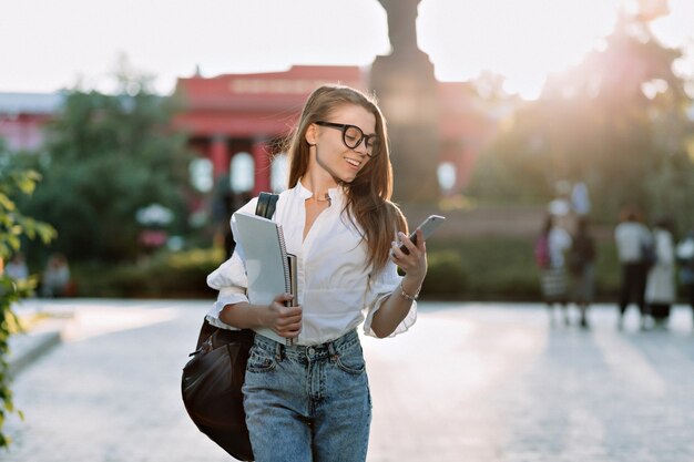 Estudante morena sorridente de óculos e camisa caminhando ao ar livre com laptop e mochila e olhando no smartphone à luz do sol