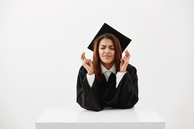 Estudante nervosa na esperança de obter seu diploma, com os dedos cruzados sobre uma parede branca