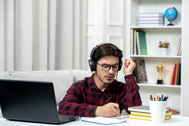 Estudante on-line bonitinho de camisa xadrez com óculos estudando no computador confuso e escrevendo