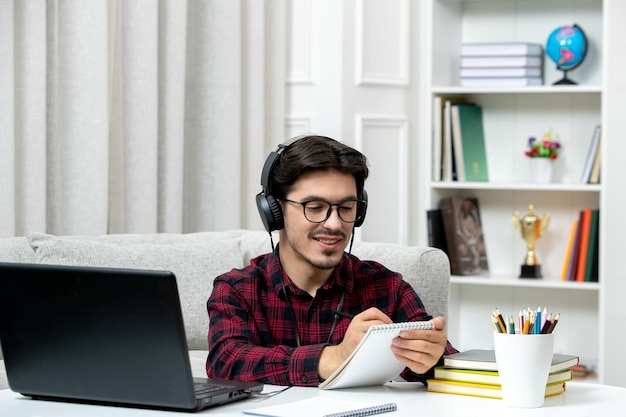 Estudante on-line bonitinho de camisa xadrez com óculos estudando no computador escrevendo notas