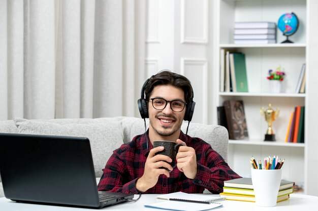 Estudante on-line bonitinho de camisa xadrez com óculos estudando no computador sorrindo e feliz
