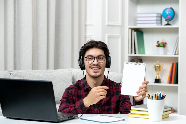 Estudante on-line bonitinho de camisa xadrez com óculos estudando no computador sorrindo segurando notas