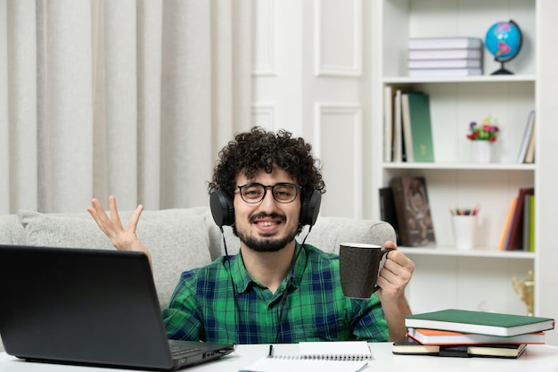 Estudante on-line bonito jovem estudando no computador em óculos de camisa verde com copo feliz