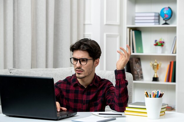 Estudante online jovem de camisa xadrez com óculos estudando no computador confuso