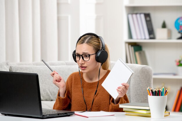 Estudante online linda garota de óculos e suéter estudando no computador segurando caneta e bloco de notas