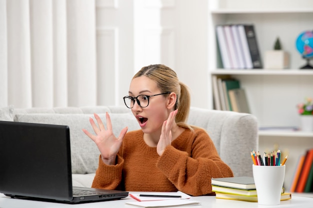 Estudante online linda garota de óculos e suéter laranja estudando no computador chocado