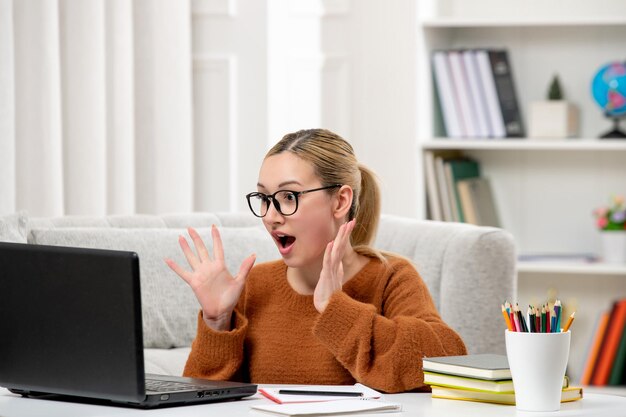 Estudante online linda garota de óculos e suéter laranja estudando no computador chocado