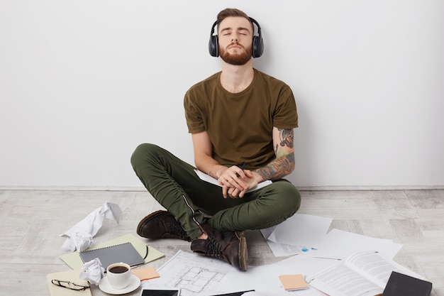 Estudante relaxado e moderno em roupa casual, senta-se no chão de pernas cruzadas, curte rock em fones de ouvido,