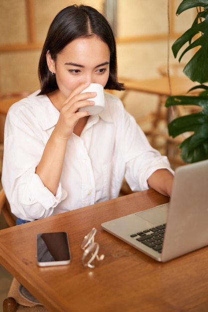 Estudante sentado no café com uma xícara de café jovem mulher asiática trabalhando no laptop no restaurante sentado