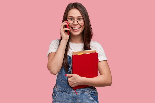 Estudante sorridente conversa ao telefone durante o intervalo, segura um celular moderno, usa macacão jeans, segura um bloco de notas espiral