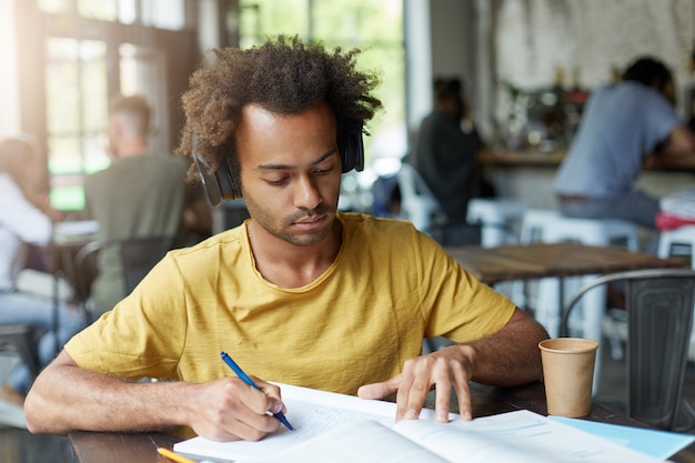 Estudante universitário afro-americano elegante fazendo lição de francês no refeitório, estudando pronúncia e ortografia, ouvindo tarefas de áudio usando fones de ouvido enquanto aprende novas palavras