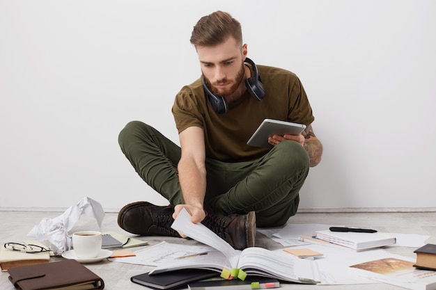 Estudante universitário do sexo masculino, urso e preocupado, com um penteado moderno, olha atentamente para o livro e segura um tablet moderno