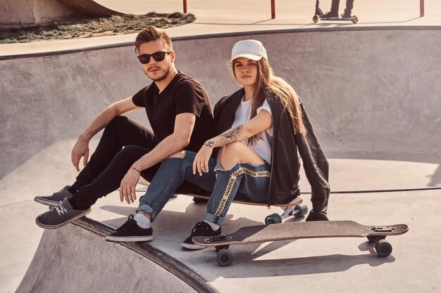 Estudantes da moda estão sentados no ensolarado skatepark com seus longboards. Há outros skatistas ao fundo.