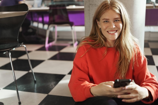 Estudantes de trabalho de meio período e conceito de estilo de vida closeup de mulher ruiva atraente feliz sente-se cantina universitária de café no chão sorrindo câmera usando telefone celular mandando mensagens de texto para amigos assistindo vídeos