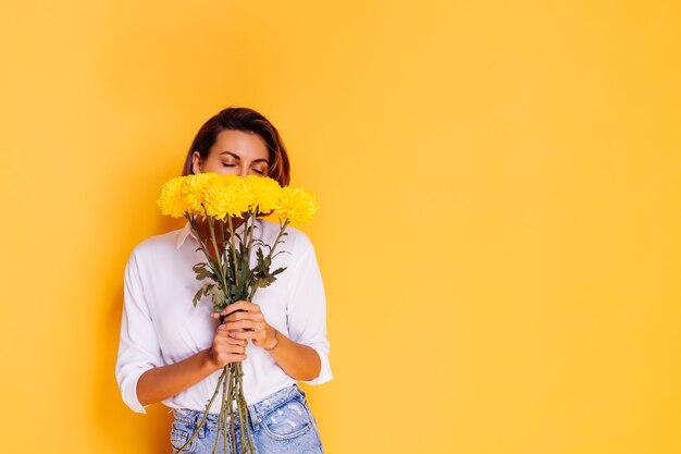 Estúdio, tiro em fundo amarelo. Mulher caucasiana feliz, cabelo curto, vestindo roupas casuais, camisa branca e calça jeans, segurando um buquê de ásteres amarelos