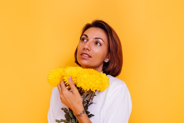 Estúdio, tiro em fundo amarelo. Mulher caucasiana feliz, cabelo curto, vestindo roupas casuais, camisa branca e calça jeans, segurando um buquê de ásteres amarelos