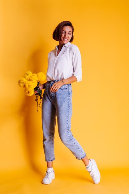 Estúdio, tiro em fundo amarelo. Mulher caucasiana feliz, cabelo curto, vestindo roupas casuais, camisa branca e calça jeans, segurando um buquê de ásteres amarelos