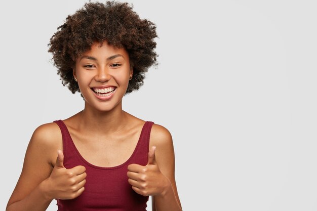 Eu digo sim para novas oportunidades. Jovem negra alegre com penteado afro, puxando as mãos com o polegar para cima