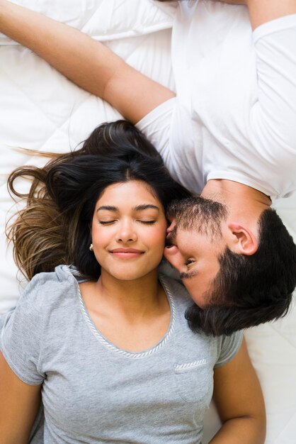 Eu te amo! Amar o jovem em pjs beijando a bochecha de sua namorada feliz e sorridente enquanto estava deitado juntos na cama