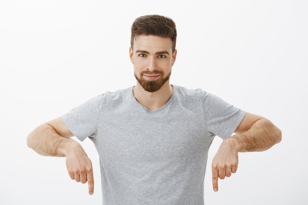 Foto grátis eu tenho que comprar. homem confiante, entusiasmado e carismático criativo, com barba e corte de cabelo castanho apontando para baixo com os braços erguidos, sorrindo com um olhar entusiasmado e encantado garantindo ao amigo que experimente uma parede cinza