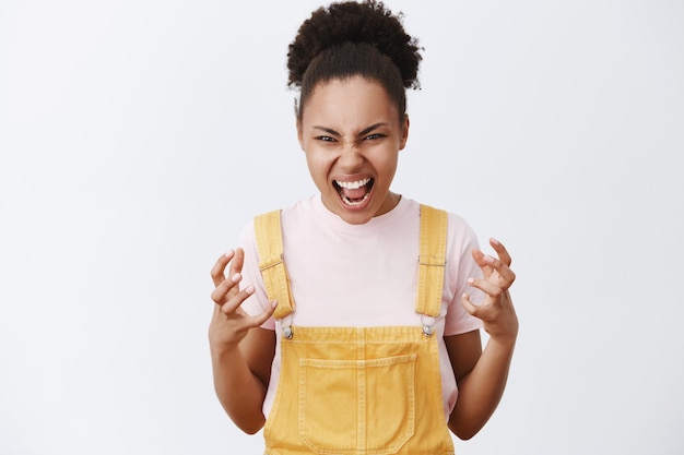 Eu vou te sufocar com as mãos nuas. Retrato de uma mulher afro-americana emotiva e irritada com um macacão amarelo da moda, gesticulando com as palmas das mãos de ódio, gritando de raiva sobre a parede cinza