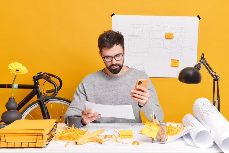 Foto grátis europeu barbudo sério concentrado no papel usa telefone celular, faz desenhos para construtora rodeados de papel e adesivos de rolos de planta. conceito de trabalho.