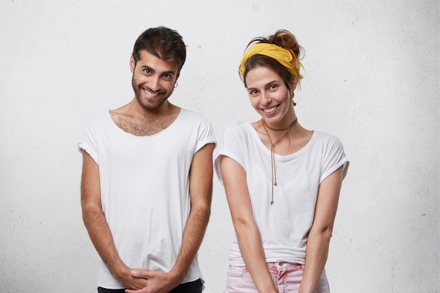 Foto grátis europeu masculino e feminino vestindo camiseta branca, sorrindo sinceramente com expressões tímidas. pessoas positivas demonstrando bom humor e sorrisos