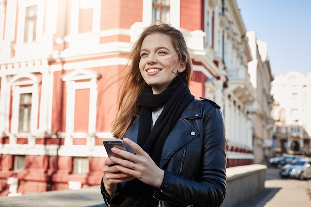 Excelente dia para aventuras. Retrato da cidade de mulher atraente europeu, andando na rua, segurando o smartphone