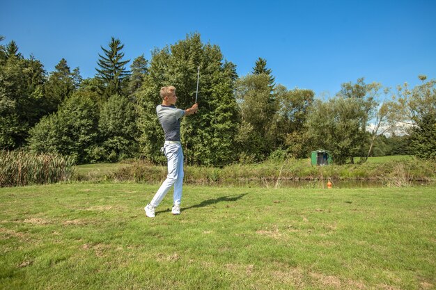 Excelente foto de um jovem jogando golfe em um campo cercado por árvores em um dia ensolarado