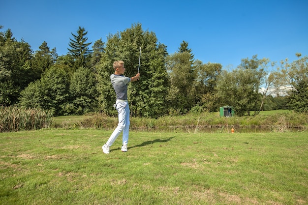 Excelente foto de um jovem jogando golfe em um campo cercado por árvores em um dia ensolarado