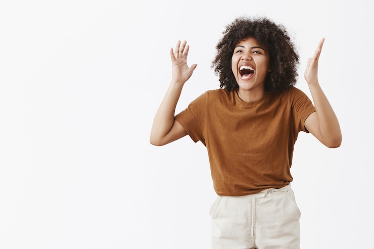 Foto grátis excessivamente emotiva, despreocupada e feliz, elegante mulher afro-americana moderna em uma camisa marrom da moda gesticulando com as mãos levantadas perto da cabeça e rindo alto olhando para a esquerda