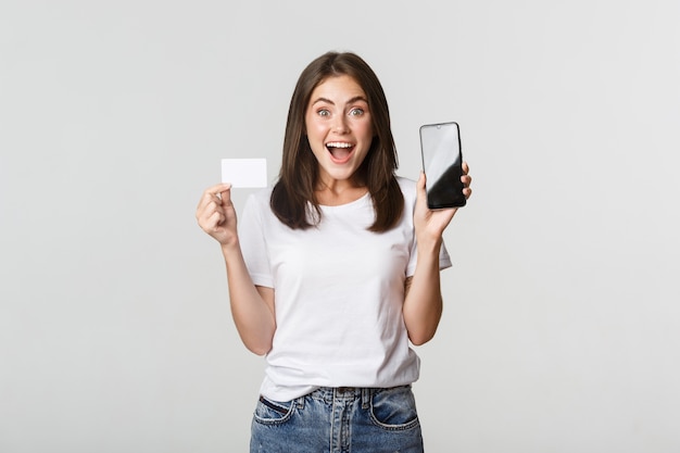 Excitada e surpresa linda garota mostrando cartão de crédito e aplicativo de banco de celular na tela.