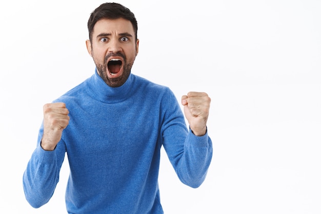 Excitado, jovem intenso de aparência séria apostou em uma partida de esporte de futebol, cerrou os punhos e gritou torcendo pelo time, gritando como se estivesse olhando para a tela da TV, quer que o time marque um gol, parede branca