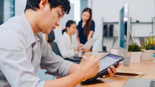 Executivo bem sucedido Ásia jovem empresário inteligente casual vestir desenho, escrever e usar caneta com tablet digital pensando em idéias de busca de inspiração, trabalhando o processo no escritório moderno.