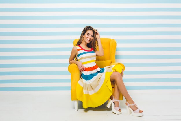 Expressando brilhantes emoções positivas de alegre jovem elegante em vestido colorido, se divertindo na cadeira amarela na parede branca azul listrada. Horário de verão, alegria, sorriso, felicidade.