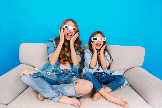 Expressando emoções verdadeiras felizes loucas para a câmera da mãe elegante e a filha em roupas jeans no sofá isolado sobre fundo azul. Usando óculos 3D, se divertindo juntos