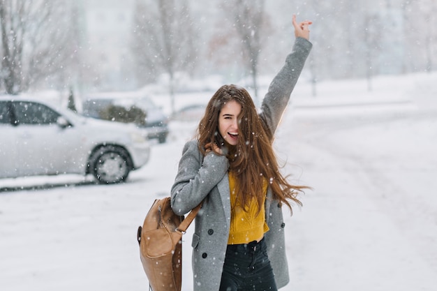 Expressando felizes emoções verdadeiras positivas de mulher andando em tempo nevando no inverno na rua. Mulher incrível e animada com longos cabelos castanhos, desfrutando da queda de neve, se divertindo.