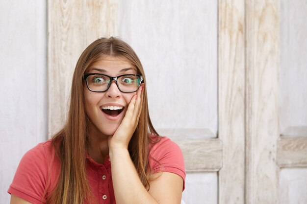 Expressões e emoções do rosto humano. retrato de uma jovem funcionária atônita, vestindo camisa pólo e óculos retangulares, segurando a mão na bochecha e abrindo a boca amplamente